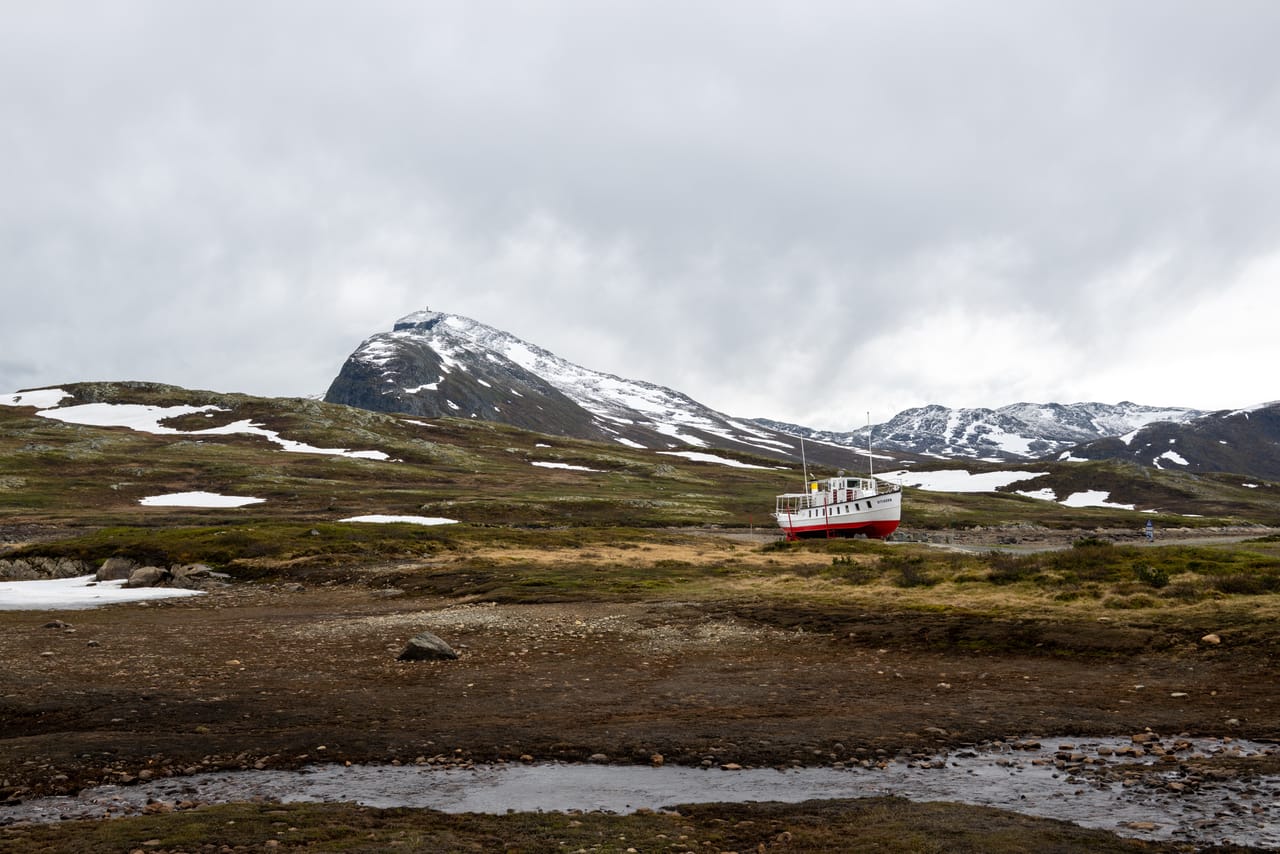 MB Bitihorn på land ved Bygdin, med fjellet Bitihorn (1607 moh) og en bekk i forgrunnen.