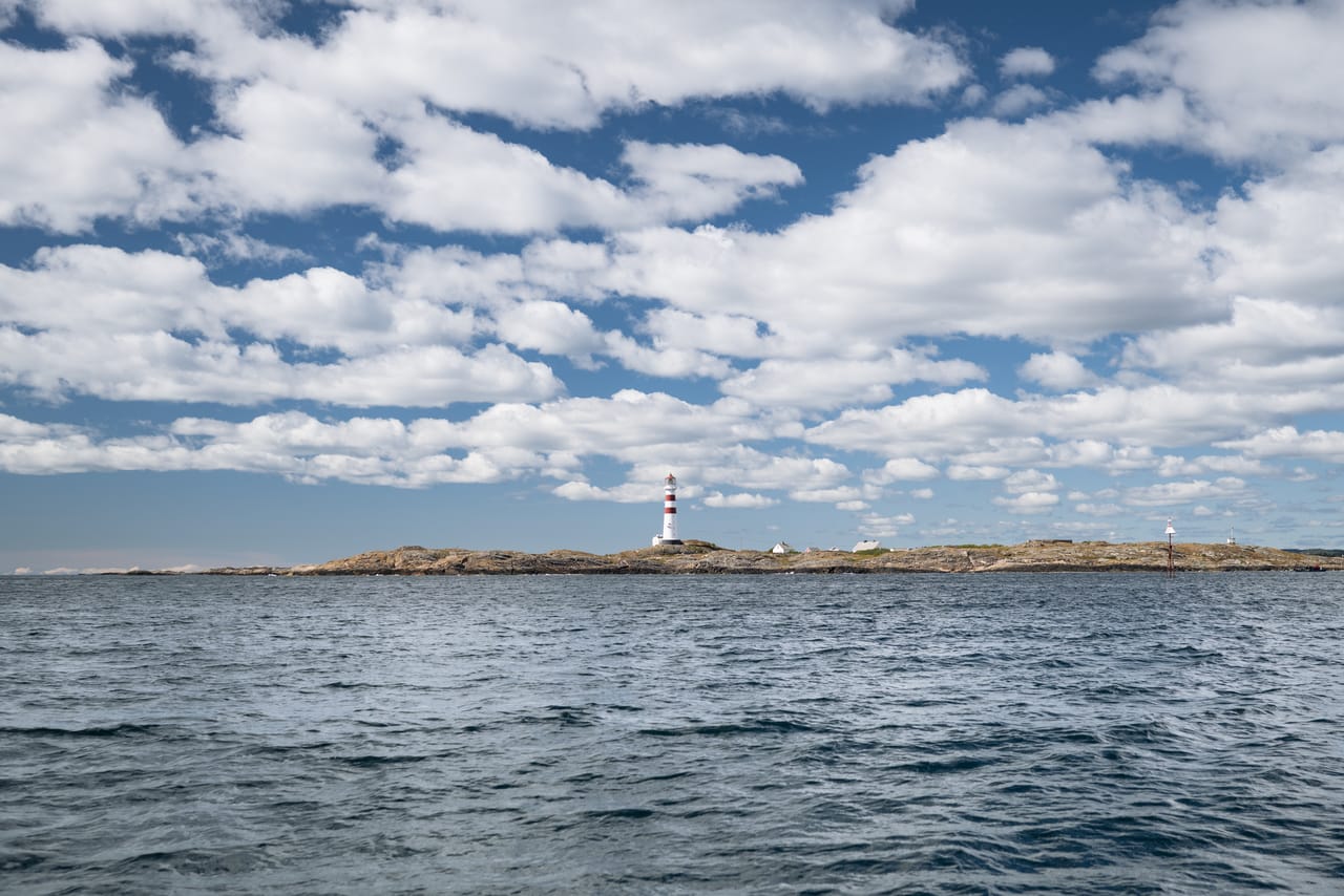 Oksøy fyr sett på avstand fra sjøen, det rødstripete tårnet omgitt av lave holmer.