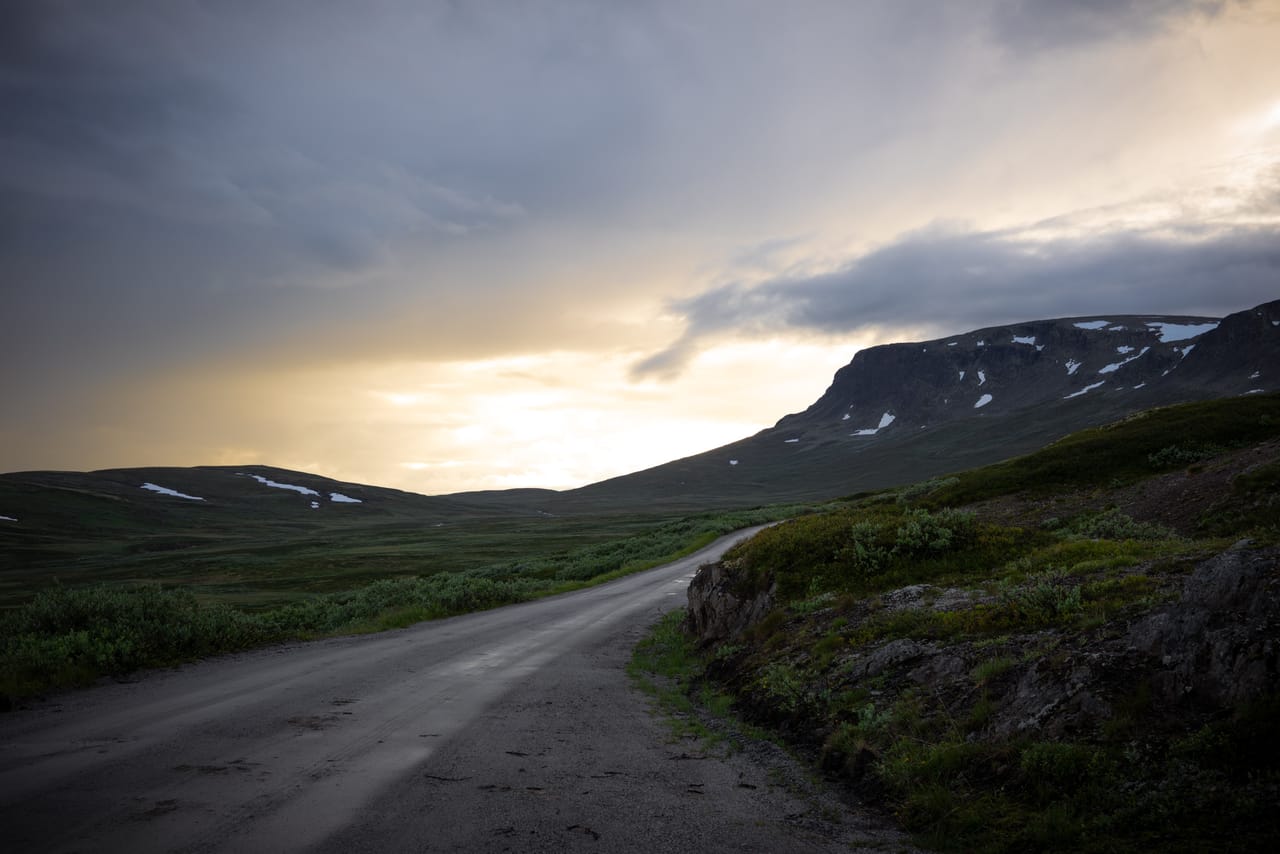 Grusveien opp mot Hallingskarvet i kveldslys, med et bratt fjellparti og snøflekker mot en fargerik himmel.