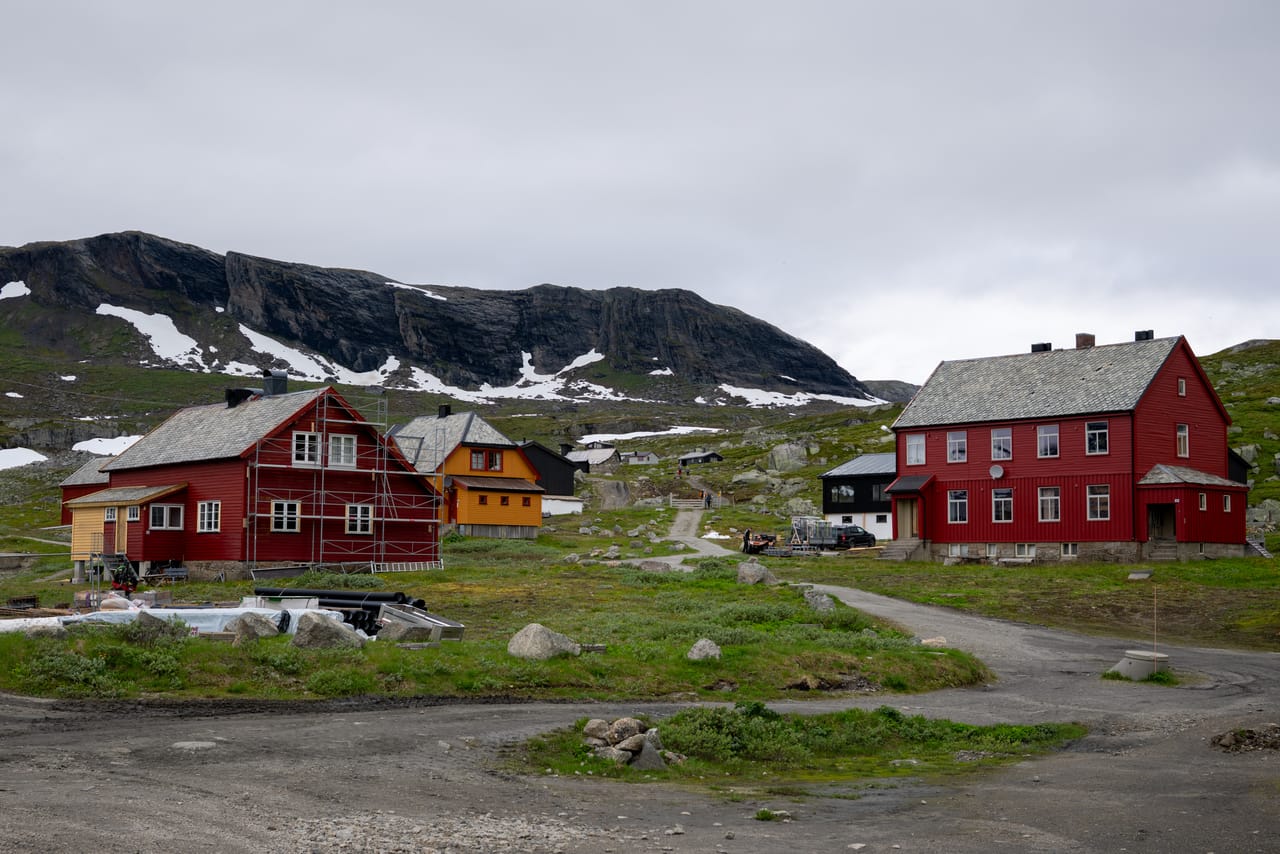 Bygninger ved Finse under oppussing, med et bratt fjellparti og snøflekker i bakgrunnen.
