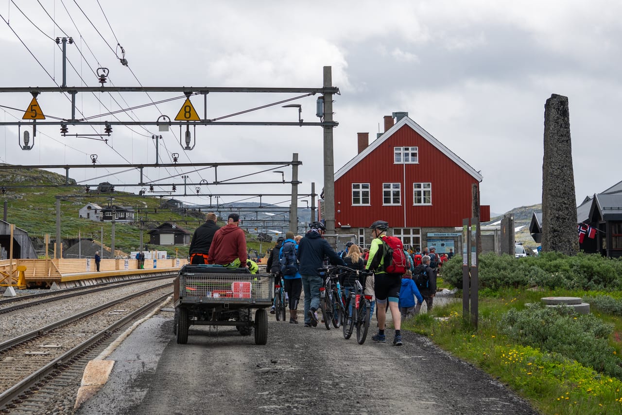 Turgåere og syklister ved Finse stasjon, med den røde stasjonsbygningen og togskinner i forgrunnen.