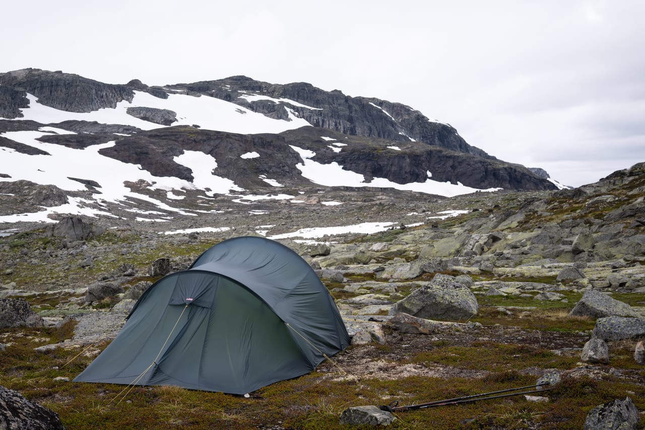 Telt satt opp mellom steinene i et værhardt landskap, med et snødekt fjell i bakgrunnen.