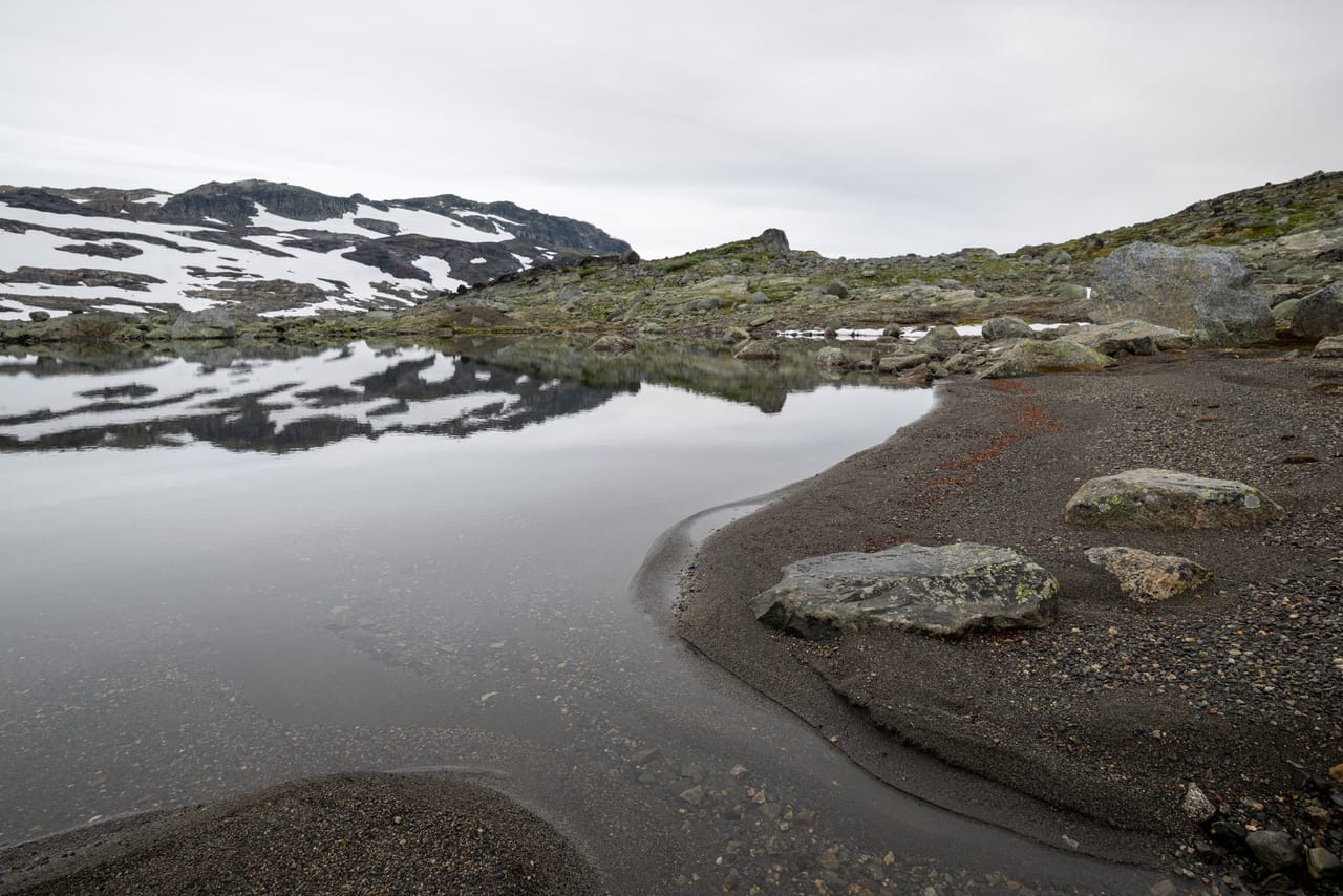 Stille tjern som speiler fjellet, med en steinete strand i forgrunnen.
