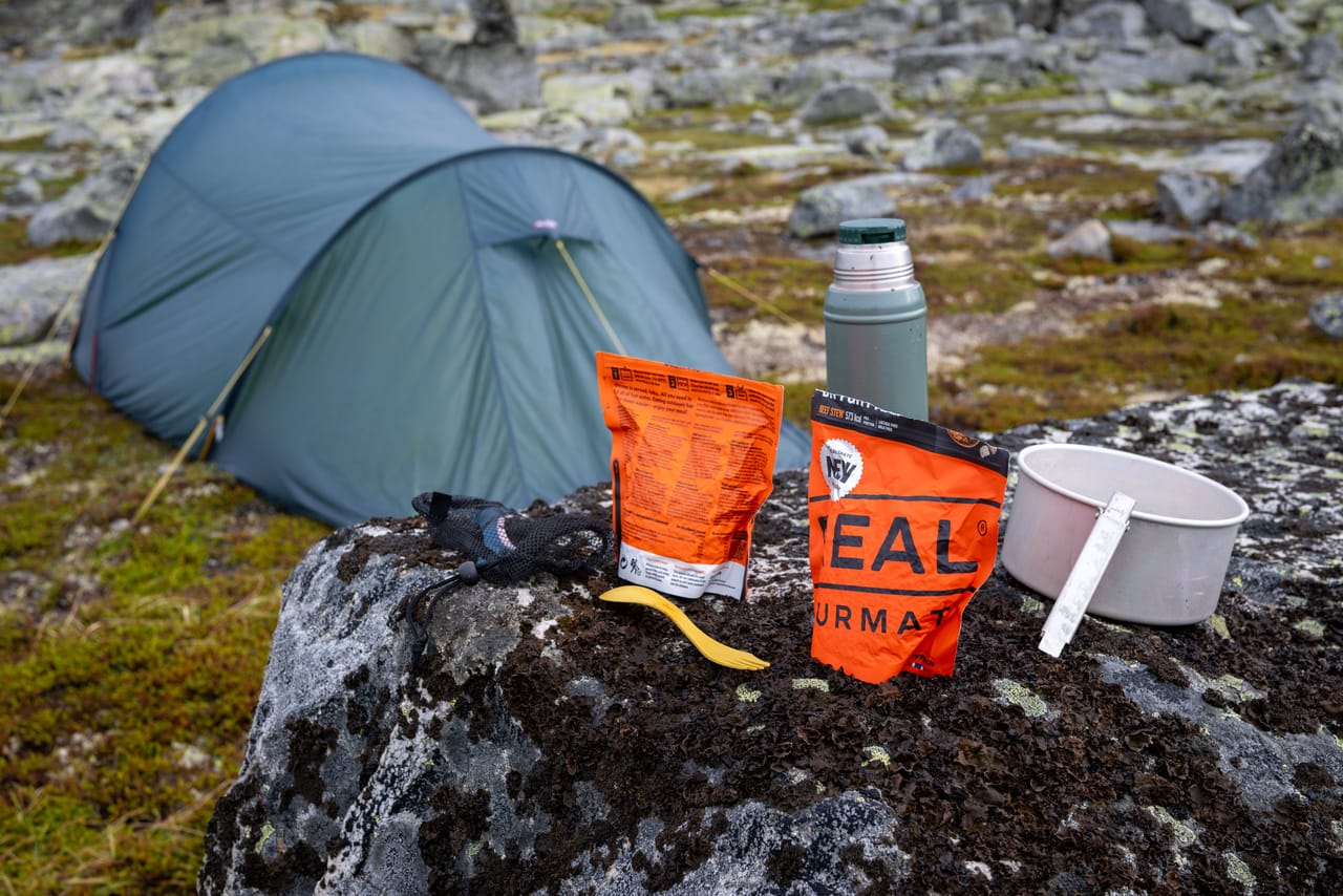 Frysetørket tursmat, kopp og termos satt fram på en stein ved teltet.