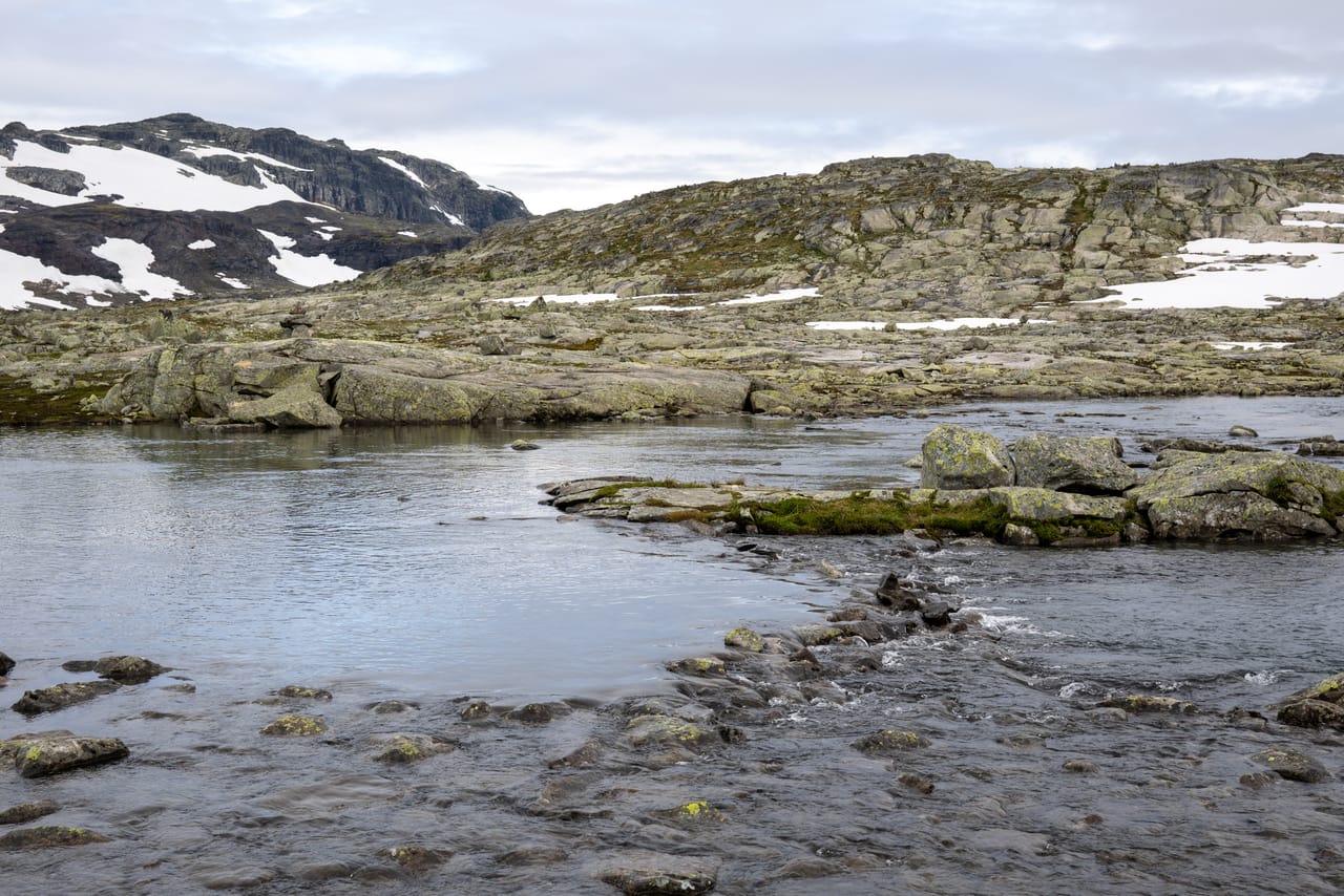 Steinete høyfjellsterreng med varde og små tjern, snøflekker i bakgrunnen.