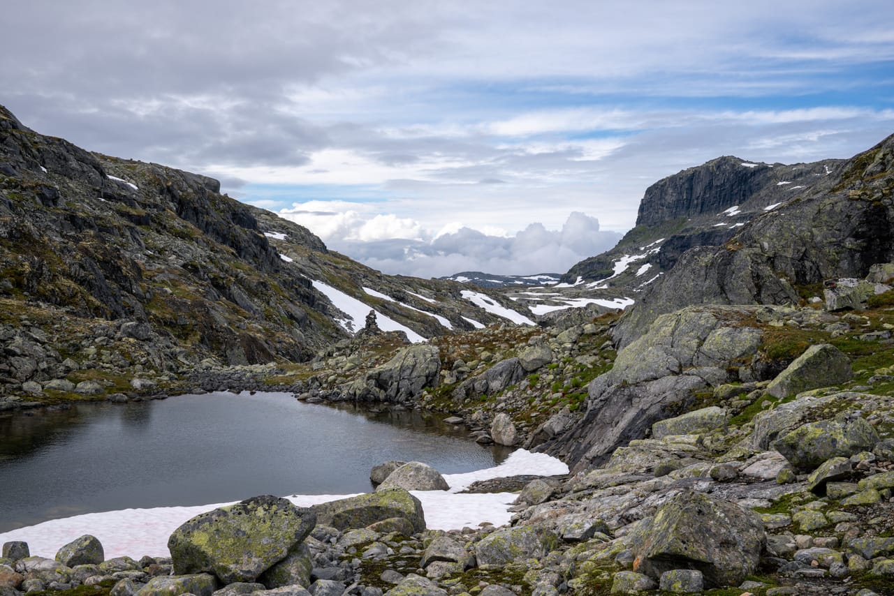 Utsikt gjennom en fjelldal med tjern, varde og snøflekker mellom bratte fjellsider.