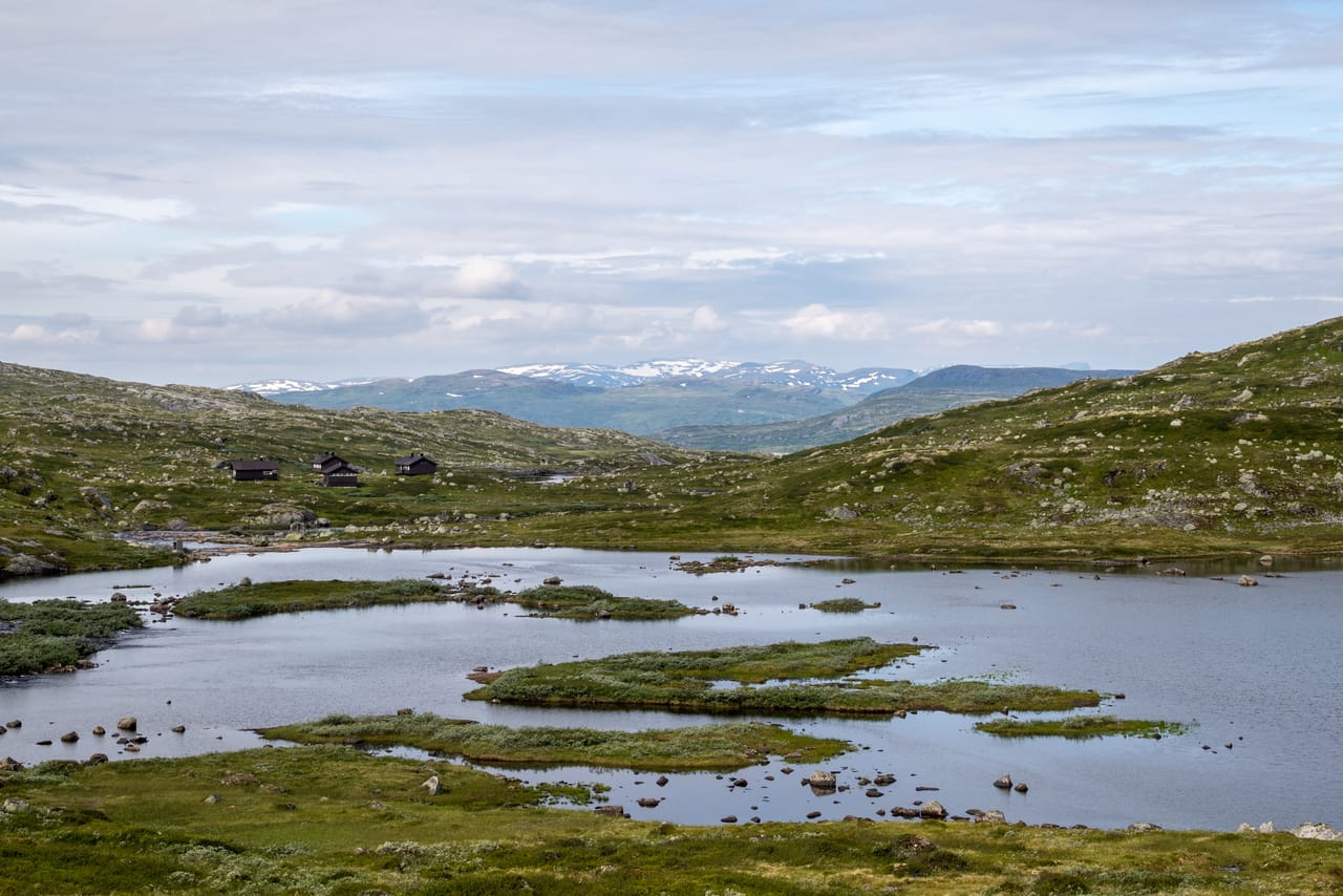 Utsikt over vidda med et tjern, Kjeldebu turisthytte i det fjerne og snødekte fjell mot horisonten.