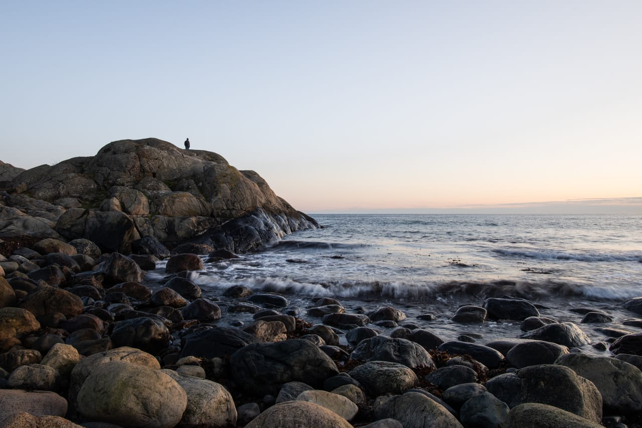 Rullesteinstrand mot åpent hav i skumringslys, med en person på et svaberg til venstre, i Raet nasjonalpark.